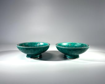 May include: Two teal-colored ceramic bowls with a pedestal base. The bowls have a textured surface and a silver rim with small white floral accents. The bowls are displayed on a white surface.