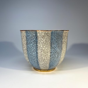 May include: A ceramic tea cup featuring a vertical striped pattern in alternating light blue and white. The surface has a distinct crackle glaze finish throughout. The rim and the base of the cup are accented with a thin, metallic gold-colored trim.
