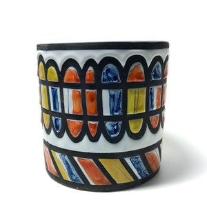 May include: A cylindrical ceramic vase with a black rim and base. The white body features a repeating pattern of colourful rectangles and arches in yellow, orange, and blue, outlined in black. The vase has a modern, geometric design.