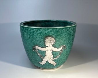 May include: A green ceramic planter with a white silhouette of a child holding a garland. The planter has a speckled glaze.