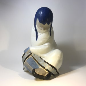 Könnte beinhalten: Keramikfigur einer sitzenden Figur mit blauen Haaren und einem weißen Gewand. Die Figur trägt einen gemusterten blauen und grauen Rock und schwarze Schuhe. Ruhiger Gesichtsausdruck.