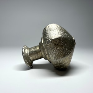 May include: A silver metal top with a rounded, textured design. The top has a cylindrical base with a raised rim.