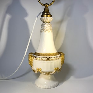 May include: A white porcelain table lamp with gold accents and a decorative design. The lamp has a round base and a tall, slender body with a gold band around the middle. The lamp is decorated with a gold leaf pattern and has a gold-colored cord.