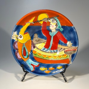 Puede incluir: Un plato de cerámica colorido con una ilustración caprichosa de un pescador en un bote, atrapando un pez grande con una lanza. El plato tiene un fondo azul con acentos naranja, amarillo y verde. La firma "BegSimone" es visible en la parte inferior del plato.