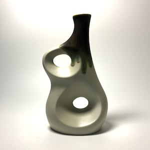 May include: White ceramic vase with a unique abstract design. The vase has two circular openings and a green and brown glaze on the neck.
