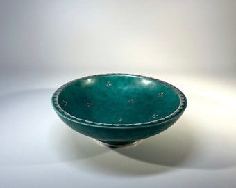 May include: A teal ceramic bowl with a silver rim and silver floral accents. The bowl has a slightly flared rim and sits on a small pedestal.