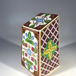May include: A ceramic box with a geometric pattern in shades of red, green, blue, and gold. The box has a square base and a rectangular top.