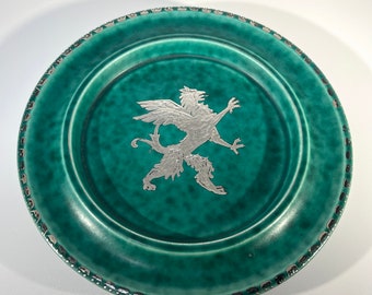 May include: A green ceramic plate with a silver-colored griffin design in the center. The plate has a decorative border.
