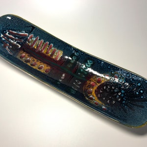 Puede incluir: Un plato de servir de cerámica azul largo y estrecho con un diseño abstracto colorido. El diseño presenta acentos rojos, amarillos y verdes.