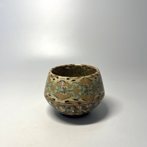 Puede incluir: Una taza pequeña de cerámica marrón con un patrón geométrico en tonos marrones, beige y verde.