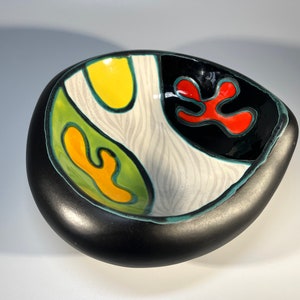Può includere: Una ciotola in ceramica nera con un design astratto unico. La ciotola presenta uno sfondo bianco con forme astratte verdi, gialle e rosse.
