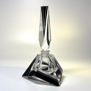 May include: A clear glass perfume bottle with a geometric design. The bottle has a faceted, triangular base with black accents and a tall, pointed stopper with black and white stripes. The bottle is empty.
