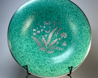 May include: A large, round, green ceramic plate with a silver floral design. The plate is on a black metal stand.
