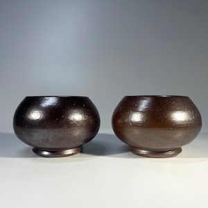 Puede incluir: Dos cuencos de cerámica marrón oscuro con forma redondeada y un borde ligeramente acampanado. Los cuencos tienen un acabado liso y brillante.