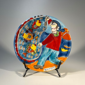 Puede incluir: Un plato redondo de cerámica con una ilustración colorida de una persona sosteniendo una red con peces. El plato tiene un fondo azul con acentos naranja, amarillo y rojo. El plato está firmado "DESIMONE" en la esquina inferior derecha.