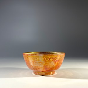 May include: A small, red and gold ceramic bowl with a decorative gold rim. The bowl has a slightly rounded shape and a smooth, glossy finish.