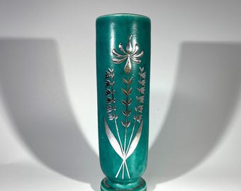May include: A teal-colored vase with silver floral designs. The vase has a cylindrical shape with a flared base and a silver flower design at the top. The silver floral designs are on the front of the vase.