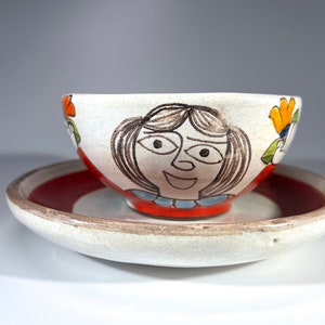 May include: A white ceramic bowl with a hand-painted design of a smiling face with brown hair and red, blue, and green accents. The bowl is sitting on a white ceramic saucer with a red band around the edge.