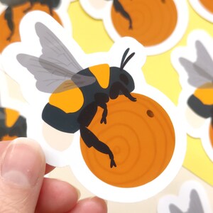 May include: A sticker of a cartoon bumblebee with black and yellow stripes, holding a round orange honeycomb. The bee has transparent wings and is on a white background.
