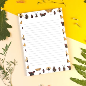 Op de afbeelding: Een wit notitieboekje met een gelinieerd interieur en een rand van kleurrijke insectenillustraties. Het notitieboekje ligt op een gele en crèmekleurige achtergrond met groene bladeren.