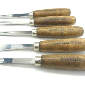 May include: A set of five wood carving tools with stainless steel blades and wooden handles. The handles are engraved with "RAMELSON USA". The tools have different blade shapes for various carving needs. The tools are arranged on a white background.