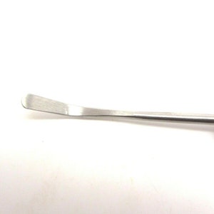 Puede incluir: Un primer plano de una herramienta de metal plateado con una cuchilla curva. La herramienta se utiliza para raspar y limpiar.