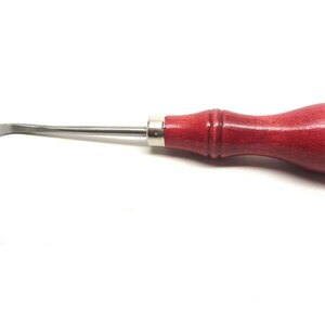 Puede incluir: Una herramienta de raspado de metal curvada con un mango de madera rojo. La herramienta se utiliza para raspar y limpiar.
