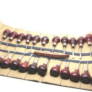 May include: A set of 24 wood carving tools with brown wooden handles and silver blades, arranged in a brown leather roll with black stitching.