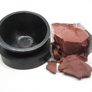 May include: A black mortar and pestle with a red clay brick inside. The clay brick is broken into pieces.