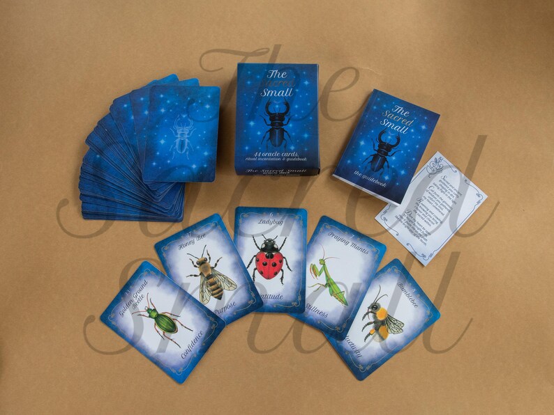 Puede incluir: Un mazo de 44 cartas or&aacute;culo titulado The Sacred Small, que incluye una caja azul y una gu&iacute;a con la ilustraci&oacute;n de un escarabajo. El conjunto contiene un mont&oacute;n de cartas azules y varias cartas individuales con insectos como mariquitas y abejas.