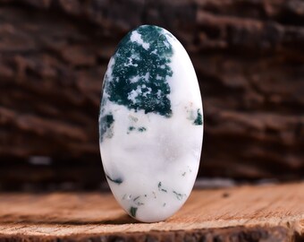Pode incluir: Cabochão oval de ágata verde e branca com um padrão manchado. A pedra está sobre uma superfície de madeira.