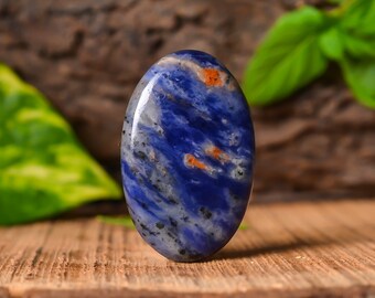 Puede incluir: Una piedra preciosa ovalada en cabujón de color azul y naranja con un patrón de mármol. La piedra está colocada sobre una superficie de madera.