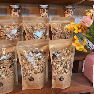 May include: A display of granola products in clear bags and jars. The bags are filled with granola and have labels with text and a logo. Jars of granola are on a shelf above the bags. A vase of flowers sits on the right.