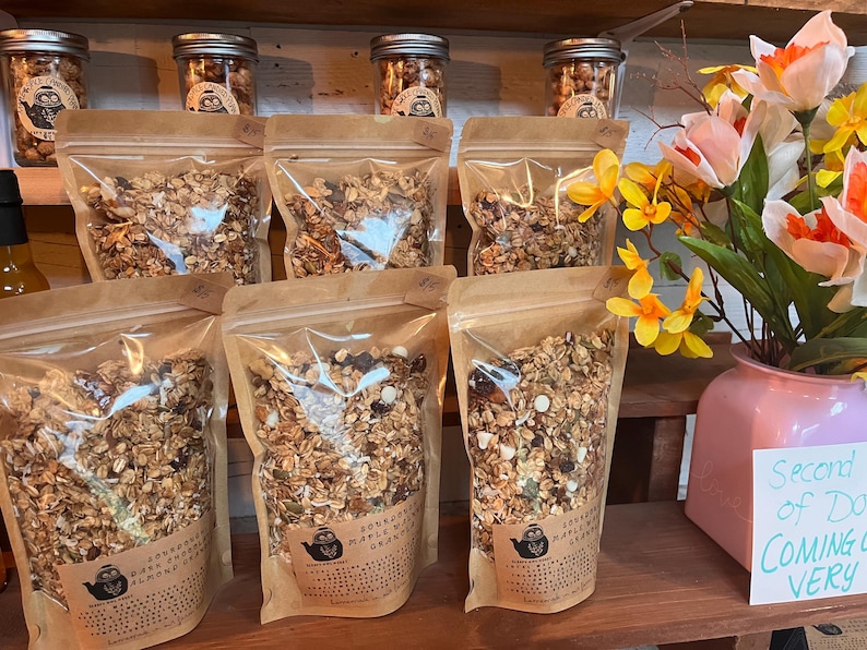 May include: A display of granola products in clear bags and jars. The bags are filled with granola and have labels with text and a logo. Jars of granola are on a shelf above the bags. A vase of flowers sits on the right.