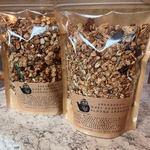 May include: Two clear plastic bags filled with granola. Each bag has a brown paper label with the text "SOURDOUGH DARK CHOCOLATE ALMOND GRANOLA" and a drawing of an owl in a teapot. The granola appears to contain oats, seeds, and dried fruit.