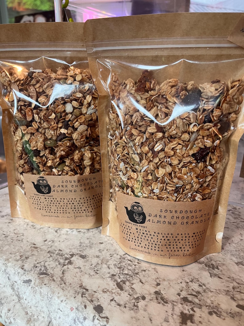 May include: Two clear plastic bags filled with granola. Each bag has a brown paper label with the text "SOURDOUGH DARK CHOCOLATE ALMOND GRANOLA" and a drawing of an owl in a teapot. The granola appears to contain oats, seeds, and dried fruit.