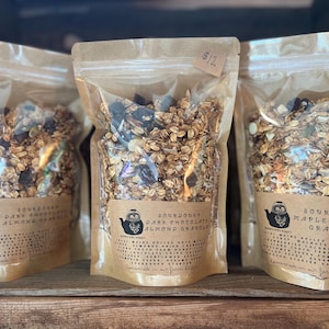 May include: Three clear plastic bags filled with granola, each with a brown label. The labels read "Sourdough Dark Chocolate Almond Granola" and "Sourdough Maple Walnut Granola". The granola appears to contain oats, nuts, and dried fruit.