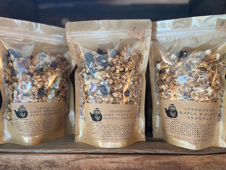May include: Three clear plastic bags filled with granola, each with a brown label. The labels read "Sourdough Dark Chocolate Almond Granola" and "Sourdough Maple Walnut Granola". The granola appears to contain oats, nuts, and dried fruit.