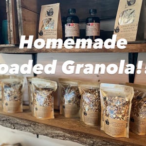 May include: A display of homemade granola and herbal tea products. Clear bags of granola are arranged on a wooden shelf, alongside bottles and tea packages. The text "Homemade Loaded Granola!!!" is displayed across the image.