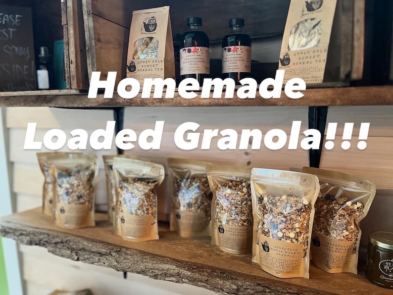 May include: A display of homemade granola and herbal tea products. Clear bags of granola are arranged on a wooden shelf, alongside bottles and tea packages. The text "Homemade Loaded Granola!!!" is displayed across the image.