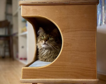 May include: A wooden cat bed with a rounded opening for the cat to enter. The bed is on four legs and has a light brown finish. A cat is peeking out of the opening.