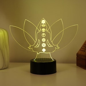 以下が含まれることがあります： 蓮の花のポーズで瞑想する人の形をした、3Dプリントされたアクリル製のランプ。ランプは暖色系の黄色い光で点灯し、7つのチャクラを照らしています。ランプは黒い台座の上に置かれています。