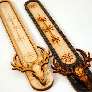 Op de afbeelding: Twee houten wierookstokjes met een herten schedel ontwerp. Een stokje is van natuurlijk hout met een lichtbruine afwerking en de andere is zwart geverfd. Beide stokjes zijn gegraveerd met runen en symbolen.