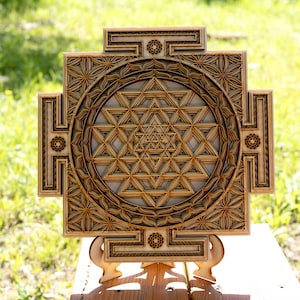 May include: A wooden wall hanging with a geometric design, featuring a Sri Yantra, a sacred symbol in Hinduism. The design is intricate and detailed, with a circular pattern of triangles and squares. The wall hanging is mounted on a small wooden stand.