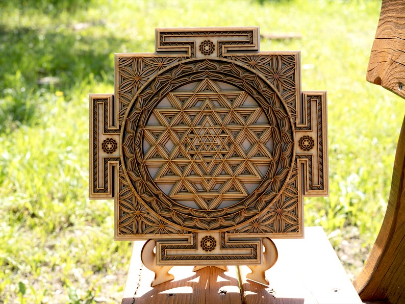 May include: A wooden wall hanging with a geometric design, featuring a Sri Yantra, a sacred symbol in Hinduism. The design is intricate and detailed, with a circular pattern of triangles and squares. The wall hanging is mounted on a small wooden stand.