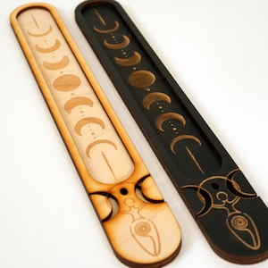 May include: Two wooden incense holders with a moon phase design. One is natural wood with a gold moon phase design, the other is black with a gold moon phase design.