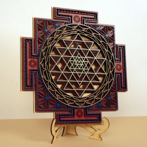 May include: A wooden Sri Yantra mandala with intricate laser-cut details. The mandala is painted in shades of red, blue, and gold. It is displayed on a wooden stand.