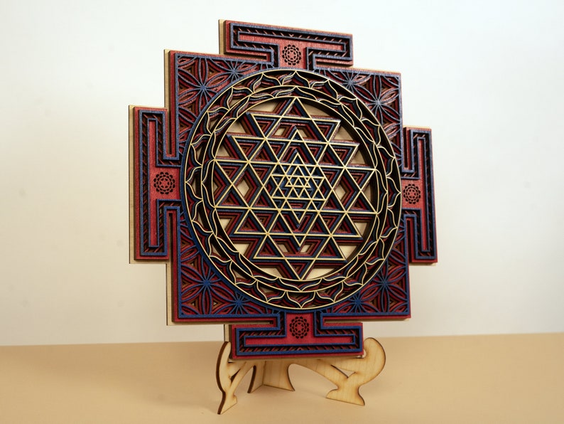 May include: A wooden Sri Yantra mandala with intricate laser-cut details. The mandala is painted in shades of red, blue, and gold. It is displayed on a wooden stand.