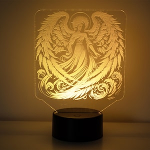 Puede incluir: Un ángel blanco con alas, grabado en un panel de acrílico transparente, iluminado desde atrás por una luz amarilla cálida. El ángel está de pie sobre un diseño de nube en espiral. El panel está montado sobre una base negra.