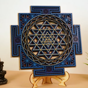 May include: A blue and gold Sri Yantra mandala with intricate geometric patterns, laser cut from wood and mounted on a wooden stand.
